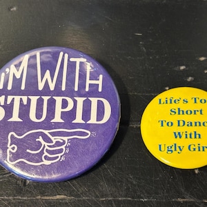 May include: Two round buttons. One is purple with the text "I'M WITH STUPID" and a pointing hand graphic. The other is yellow with the text "Life's Too Short To Dance With Ugly Girls".