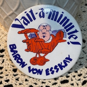 May include: A white button with the text "Wait-a-minute!" in blue, and "Baron Von Esskay" in blue. The button features a cartoon pilot in an orange airplane.