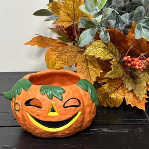May include: A ceramic jack-o'-lantern bowl with a smiling face, green leaves, and an orange body. The bowl is on a black surface, with fall foliage in the background.