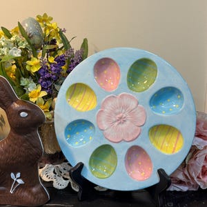 May include: A light blue ceramic Easter egg serving platter with a central pink flower and egg-shaped compartments in various colors. The eggs are decorated with stripes and dots. A chocolate bunny is on the left.
