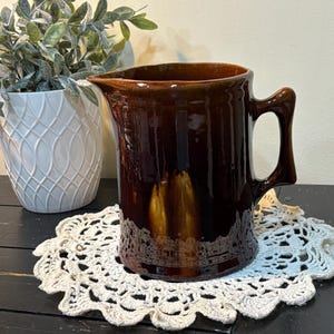 May include: A brown ceramic pitcher with a handle and spout. The pitcher is glossy and sits on a white lace doily. A white decorative pot with greenery is in the background.