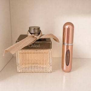 May include: A square glass perfume bottle with a ribbed texture, a metallic cap, and a beige ribbon tied around the neck, featuring the word Chloé embossed on the front. Beside it stands a tall, cylindrical rose gold refillable perfume atomizer.
