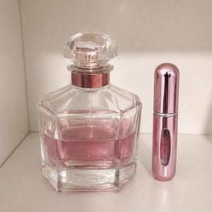 May include: A clear glass perfume bottle with a faceted hexagonal base, a pink liquid interior, and a metallic pink collar, paired with a tall, cylindrical, metallic pink travel-sized perfume atomizer with a small clear viewing window.