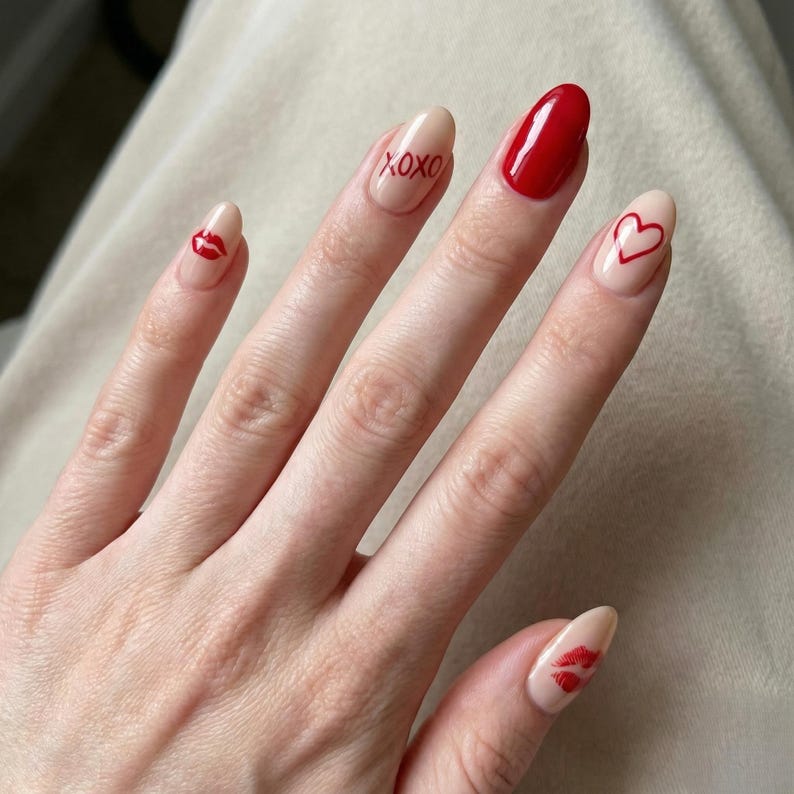 May include: A hand with almond-shaped nails, some painted with a nude base and red accents. Designs include a red heart, lips, and the text "XOXO". One nail is solid red.