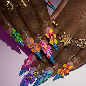 May include: A hand with long, pointed, artificial nails. Each nail is painted blue and adorned with a 3D flower design in various colors, including green, yellow, orange, and pink. Gold rings decorate the fingers.
