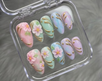 May include: A set of ten artificial nails in a clear plastic case. The nails are oval-shaped and feature a variety of pastel colors, including pink, green, blue, and purple. Each nail is decorated with a 3D flower and gold accents.