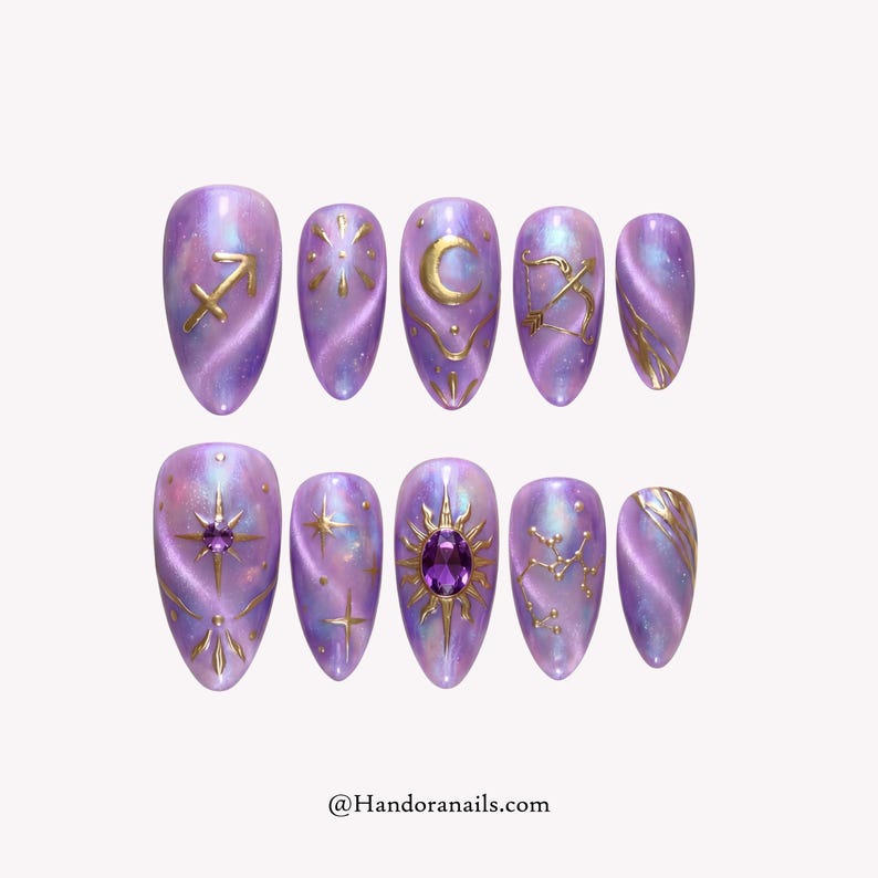 May include: Set of ten oval-shaped press-on nails in a lavender and iridescent finish. Each nail features gold celestial designs, including zodiac symbols, stars, and a sun with a purple gem. The text "@Handoranails.com" is at the bottom.