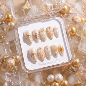 May include: A set of press-on nails in a clear plastic case. The nails are decorated with gold starfish, gold wire, floral designs, and seashell patterns. The text "Handora Nails" is visible on the bottom right. The background includes pearls and ice cubes.