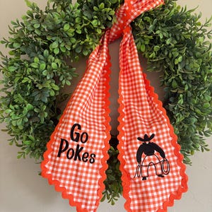 May include: A green boxwood wreath adorned with a long, orange and white gingham fabric ribbon. The ribbon features the black text "Go Pokes" on one side and a black embroidered graphic of a cowboy holding a lasso on the other side.