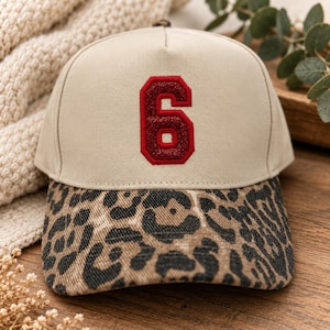 May include: A beige baseball cap featuring a red glittery number six patch centered on the front panel. The brim of the cap is decorated with a brown and black leopard print pattern. The cap has a structured crown and a curved visor.