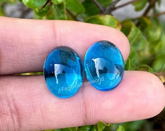 Pode incluir: Duas pedras preciosas ovais de cor azul brilhante com um acabamento liso e polido. As pedras preciosas são mantidas em uma mão, com um fundo verde frondoso.
