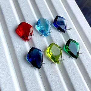 May include: Assortment of six faceted gemstones in various colours, including red, blue, green, and yellow. The gemstones are displayed on a white surface with the text "Fagemstoneworld" printed on the surface.