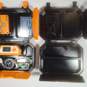 May include: Two black plastic cases with orange lids. The cases are open and show different compartments. One case contains a small circuit board, a small device with a screen, and a small device with a button. The other case contains a small circuit board and a small device with a button.