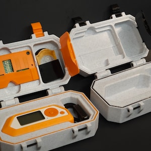 May include: Several open, textured, light grey cases with orange accents. One case contains an orange electronic device with a screen, another holds an orange circuit board, and a third has a black object. The cases are on a dark surface.