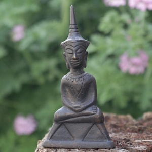 May include: A small, bronze-colored Buddha statue in a seated meditation pose. The statue has a tall, pointed headdress and is set against a blurred background of green foliage and pink flowers. The statue is approximately 4 inches tall.