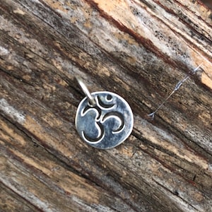 May include: A small, circular silver-toned metal pendant featuring an engraved Om symbol in the center. The pendant has a polished finish with darkened recessed areas to highlight the design and includes a small jump ring at the top for attachment.