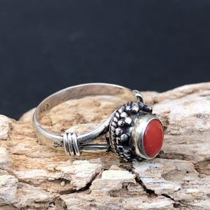 Puede incluir: Un anillo de plata con una piedra redonda de color rojo brillante. El anillo tiene un diseño detallado alrededor de la piedra y en la banda. El anillo se muestra sobre un trozo de madera flotante.