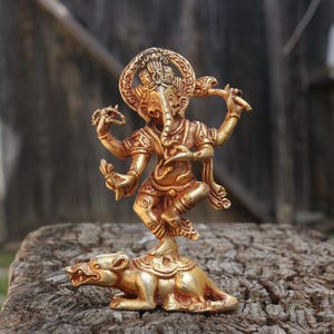 May include: A golden statue of the Hindu god Ganesha, with an elephant head and multiple arms, dancing atop a rat. The statue is detailed with intricate carvings and stands on a weathered wooden surface, showcasing a traditional religious theme.