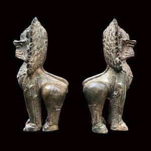 May include: Two bronze-coloured statues of mythical creatures, possibly lions or griffins, are displayed against a black background. The statues are detailed with intricate carvings and stand in a side-facing pose, showcasing their ornate manes and muscular forms.
