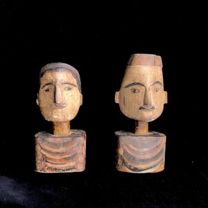May include: Two wooden figurines with painted faces and striped bodies. The figurines are carved in a simple style and have a rustic appearance.
