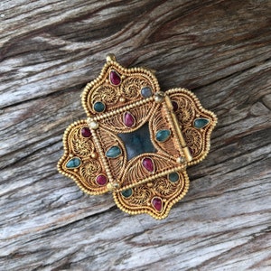 May include: Ornate gold-toned pendant with a four-leaf clover design. The pendant features intricate filigree work and is adorned with red and teal gemstones. A dark teal square is at the center of the pendant.