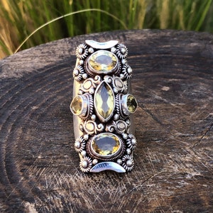 May include: A silver-toned ring featuring multiple yellow-green gemstones in various shapes. The ring has an elongated design with floral and scrollwork details. The gemstones are set in ornate bezels, creating a vintage or bohemian aesthetic.
