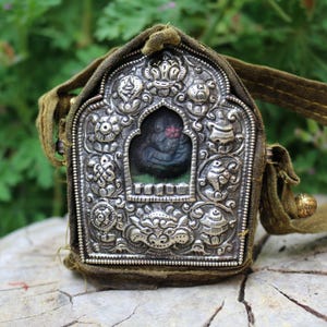 Pode incluir: Um pendente de prata ornamentado com intrincadas esculturas em relevo, apresentando uma imagem central emoldurada. O pendente está envolto numa bolsa de tecido verde com uma alça e uma pequena conta dourada. O fundo é verde desfocado.