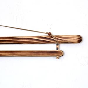 May include: A wooden poster hanger with a natural finish. The hanger has a dark brown, burnt wood appearance with a light brown cord for hanging. The design includes a small wooden bead and a lower bar for stability.