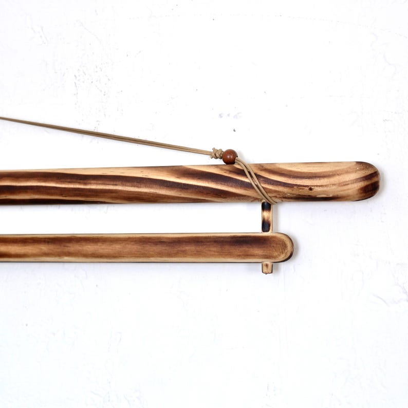 May include: A wooden poster hanger with a natural finish. The hanger has a dark brown, burnt wood appearance with a light brown cord for hanging. The design includes a small wooden bead and a lower bar for stability.