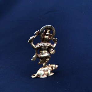 May include: A small, gold-colored statue of a deity standing on a rat. The deity has multiple arms and a halo. The statue is set against a dark blue background.