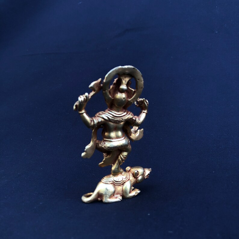 May include: A small, gold-colored statue of a deity standing on a rat. The deity has multiple arms and a halo. The statue is set against a dark blue background.