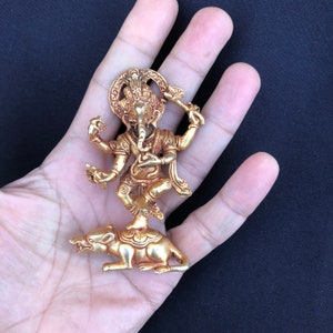 May include: A gold-colored statue of the Hindu deity Ganesha, standing on a rat. The statue has intricate details, including a crown and multiple arms. The figure is held in a hand against a dark background.