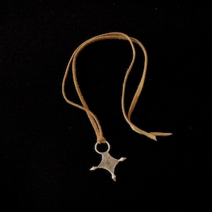 May include: A silver pendant necklace with a cross-shaped design and a tan leather cord. The pendant has a detailed pattern and is suspended from a simple ring. The necklace is displayed against a black background.