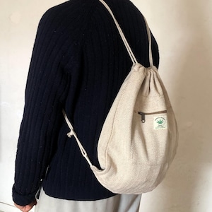 May include: Beige drawstring backpack with a herringbone pattern. The bag features a small zipped pocket and a green and white label with a leaf design. It is carried over the shoulder with white rope straps.