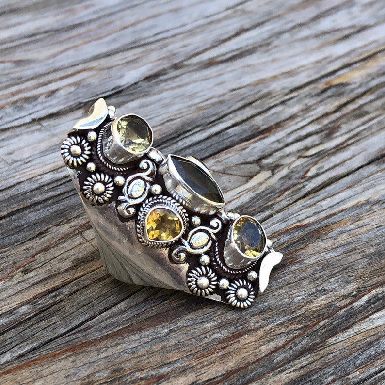 May include: A silver-toned statement ring featuring multiple gemstones in shades of yellow and green. The ring has an ornate design with floral and scroll details. The gemstones are set in various shapes, including round and oval, and are arranged across the ring's surface.