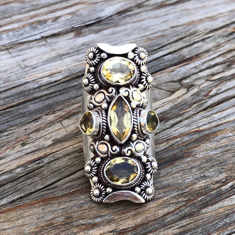 May include: A silver-toned ring featuring multiple yellow gemstones in oval and marquise shapes. The ring has a rectangular design with floral and beaded accents. The gemstones are set in a detailed, ornate setting.