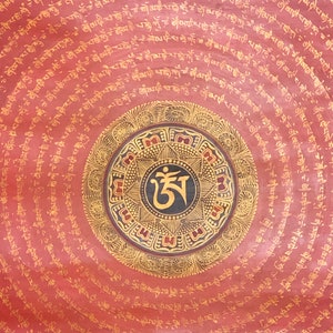 Puede incluir: Un mandala circular rojo con acentos dorados y negros. El mandala presenta un s&iacute;mbolo central con el texto "ཨ" en negro y dorado. El mandala est&aacute; rodeado de c&iacute;rculos conc&eacute;ntricos de texto en dorado.