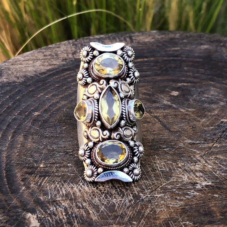 May include: A silver-toned ring featuring multiple yellow-green gemstones in various shapes. The ring has an elongated design with floral and scrollwork details. The gemstones are set in ornate bezels, creating a vintage or bohemian aesthetic.