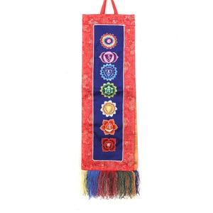 Könnte beinhalten: Ein vertikaler Wandbehang aus Stoff mit einem dunkelblauen Mittelteil, auf dem sieben Chakra-Symbole in einer Spalte eingestickt sind. Das Paneel ist von rotem Stoff mit einem dezenten Ton-in-Ton-Muster umrahmt und hat unten eine bunte Franse.