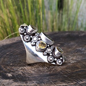 May include: A silver-toned statement ring featuring multiple gemstones in shades of green and yellow. The ring has a floral design with intricate detailing around the stones. The ring is set on a wooden surface.