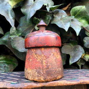 May include: A weathered wooden container with a lid, predominantly red and brown in colour. The container has a textured, aged look, and the lid is topped with a small conical finial. Green foliage forms the backdrop.