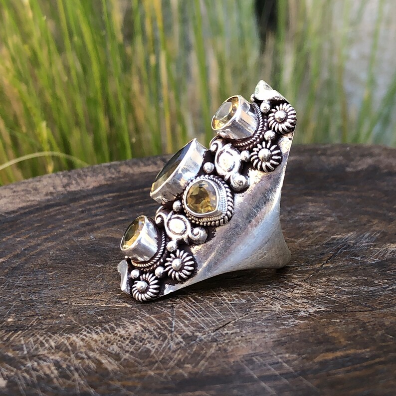 May include: A large, ornate silver ring featuring multiple yellow-green gemstones. The ring has a unique, asymmetrical design with floral and spiral embellishments. The gemstones are set in detailed, textured settings, creating a bold statement piece.
