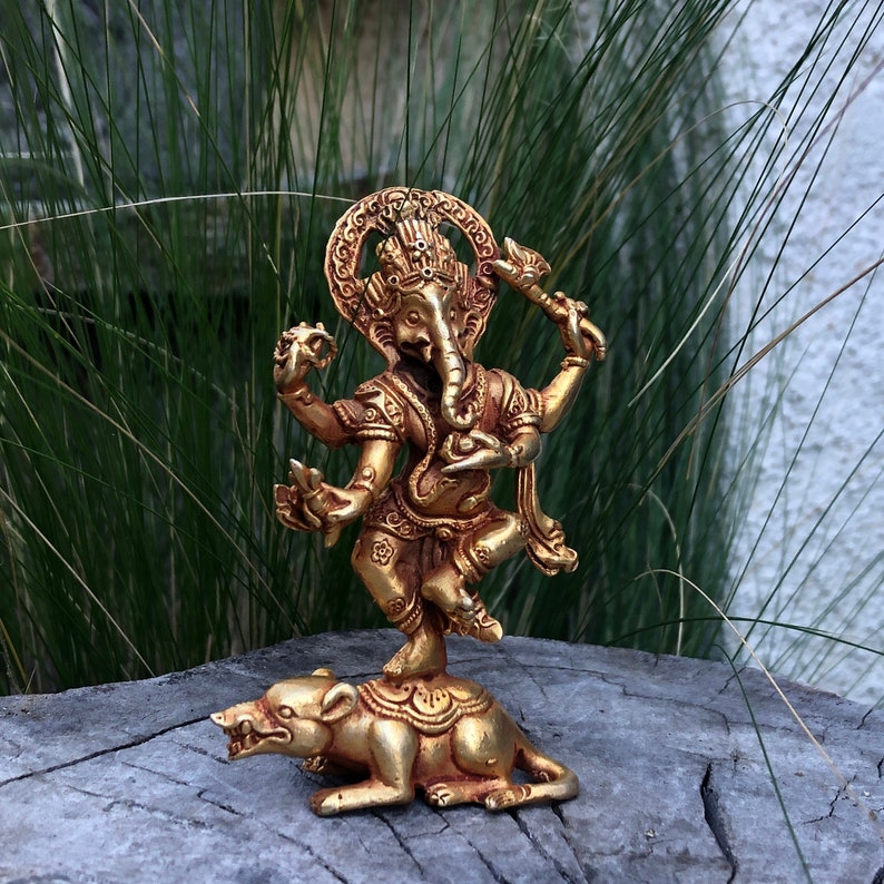 May include: A golden statue of the Hindu deity Ganesha, standing on a rat. The statue has multiple arms and intricate details. The background features green grass and a weathered wooden surface.