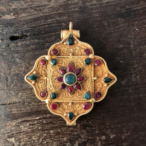 May include: Ornate gold-colored pendant with a central floral design. The pendant features a square center with a flower made of red and green gemstones. The outer edges are adorned with more red and green gemstones. The pendant has an intricate filigree pattern.