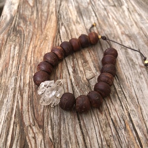 Può includere: Un braccialetto di perline di legno marrone con un ciondolo di cristallo trasparente. Il braccialetto è legato con un cordoncino marrone e ha dettagli dorati.