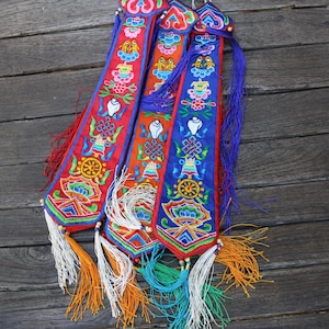 Può includere: Tre lunghi e stretti stendardi in tessuto con intricati e colorati ricami di simboli tradizionali, tra cui una ruota, un nodo e motivi floreali. Ogni stendardo è rifinito con lunghe nappe multicolori nei toni del bianco, arancione e turchese.