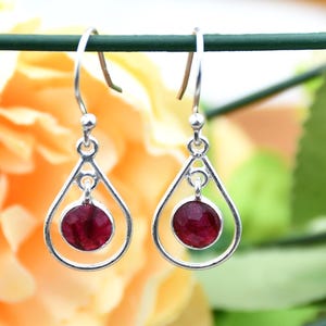 Può includere: Un paio di orecchini a goccia in argento con una gemma rotonda rosso scuro al centro di ciascun orecchino. Gli orecchini sono appesi su uno sfondo sfocato giallo e verde.