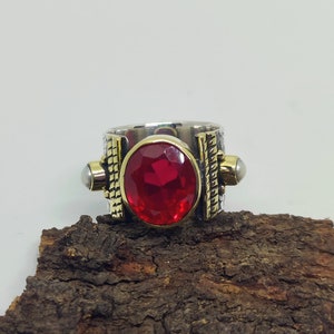 May include: A silver and gold ring with a red gemstone and two white pearls. The ring is on a piece of brown bark.