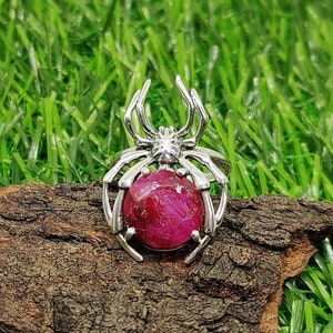 Może przedstawiać: Srebrny pierścionek w kształcie pająka z dużym, fasetowanym, rubinowo czerwonym kamieniem szlachetnym w środku.