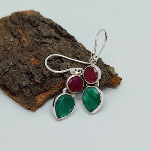 以下が含まれることがあります： 緑と赤の宝石が2つずつ付いたシルバーのドロップイヤリング。イヤリングは涙型で、上部に丸い宝石が付いています。